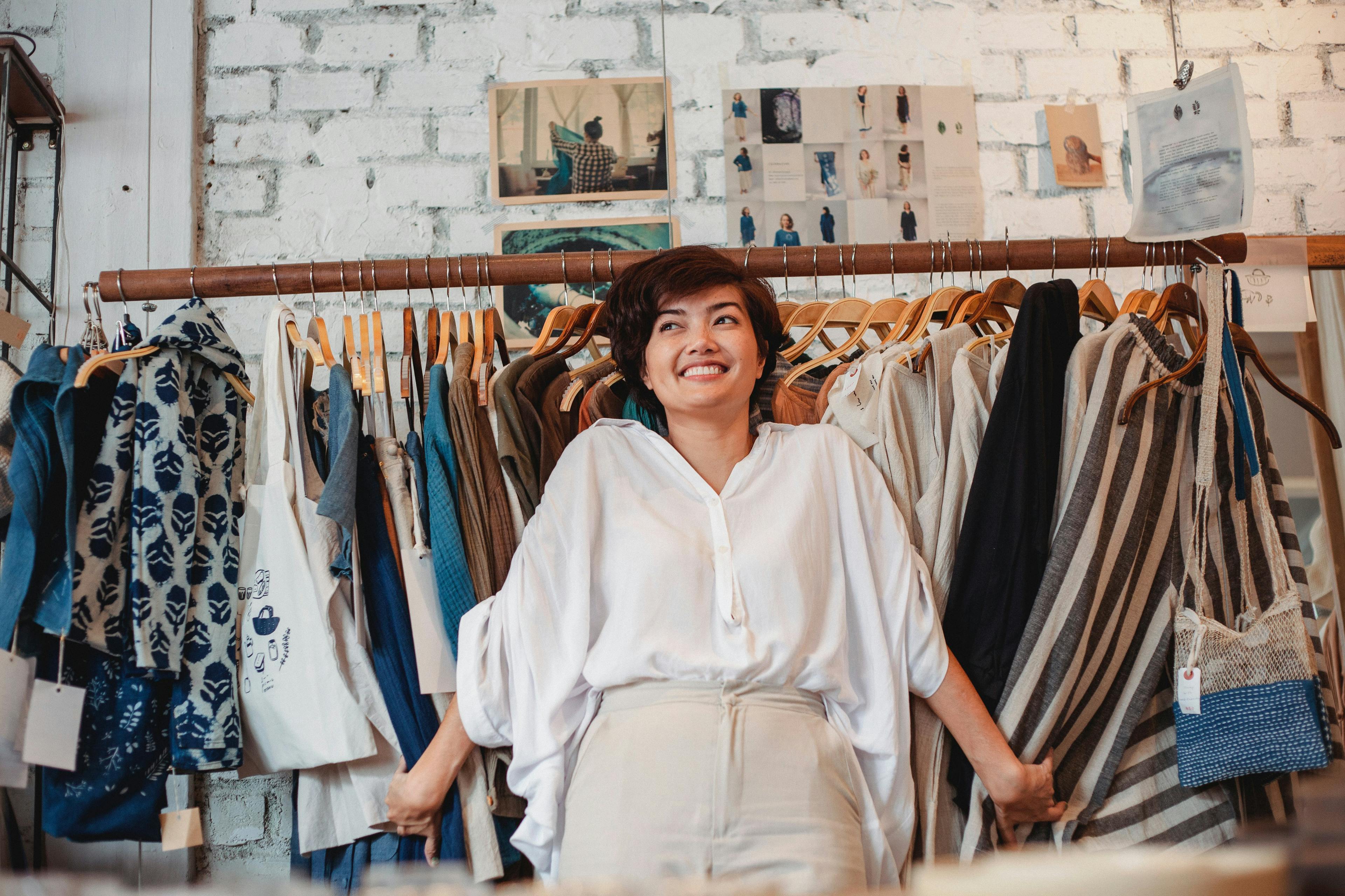 Smiling fashion boutique owner standing in front of clothing rack with various garments on display