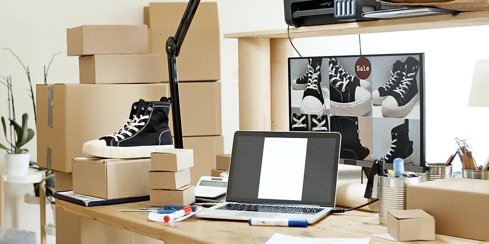 A working desk including a computer, a show, a printer and some shipment boxes.