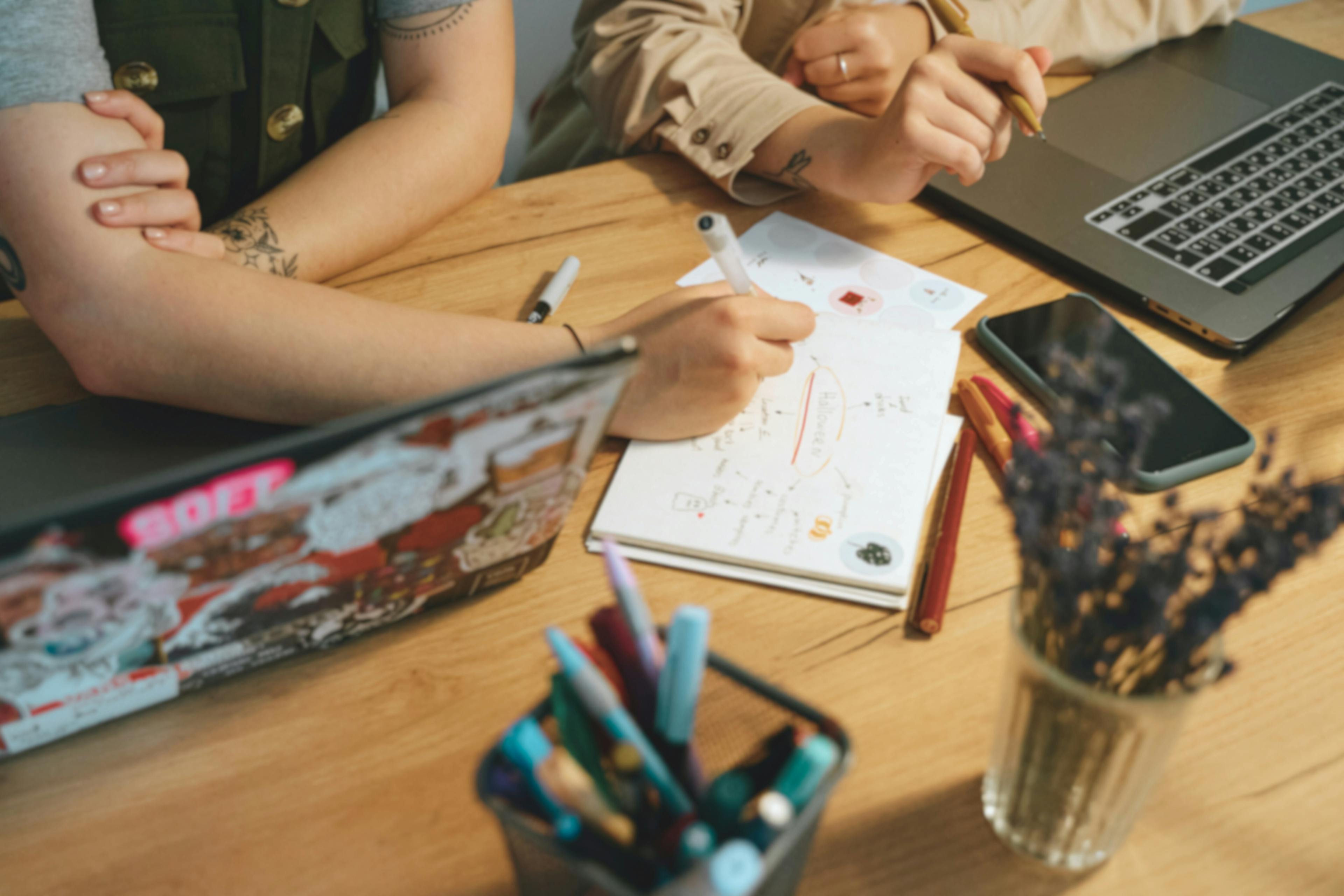 People working on creative design sketches at wooden desk with laptop and art supplies