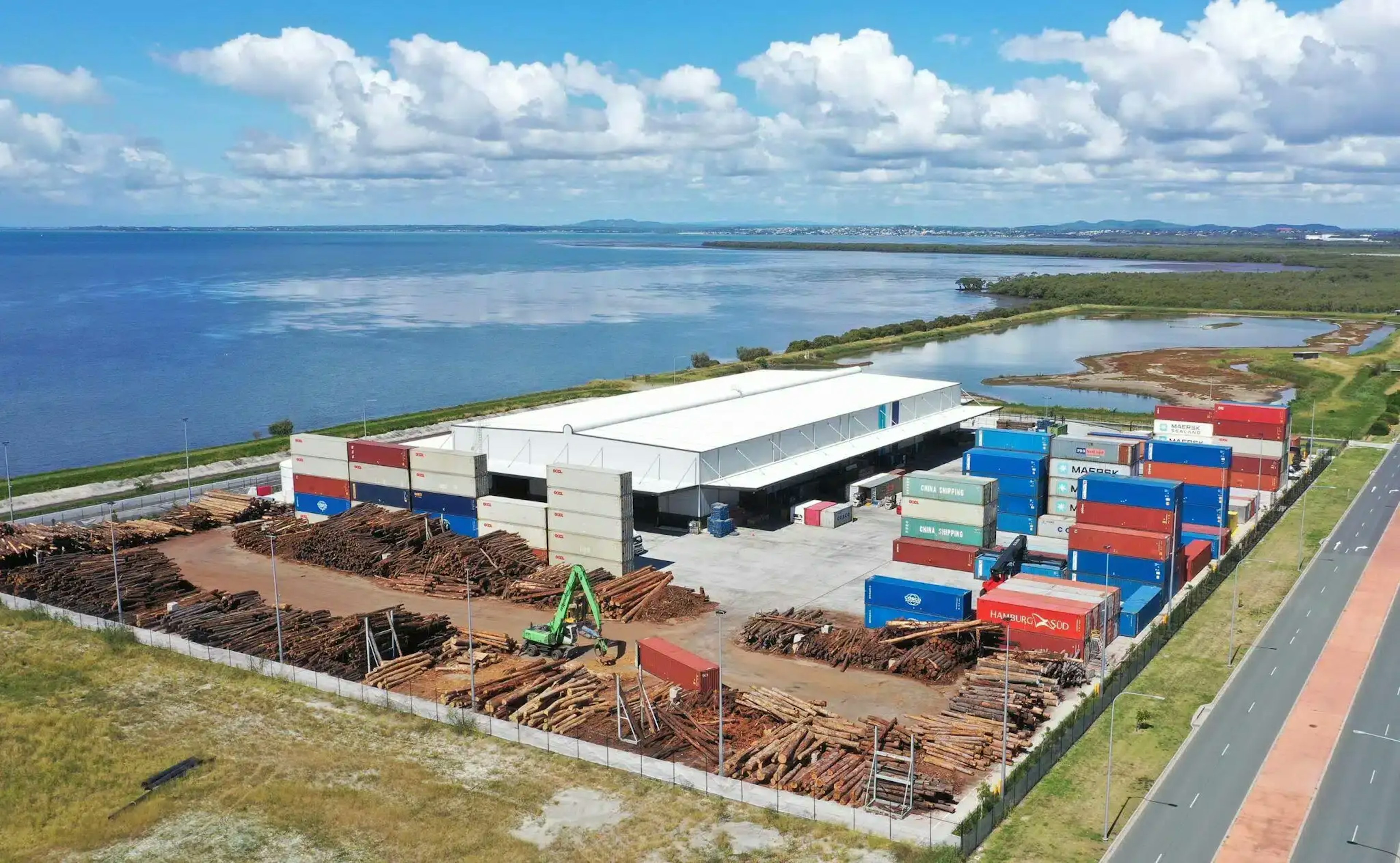Aerial view of a coastal timber processing facility with stacked logs in the yard, shipping containers, and a large white warehouse building, situated beside blue waters with distant shoreline visible under a cloudy sky