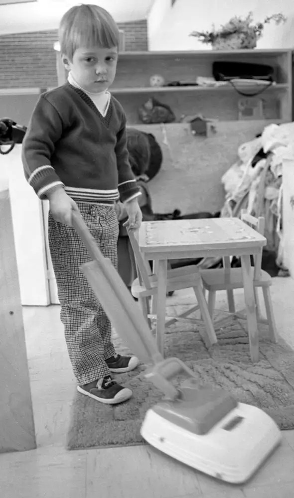 Vintage 1950s or 1960s photograph of a young kid with a serious face using a toy vacuum in a home.