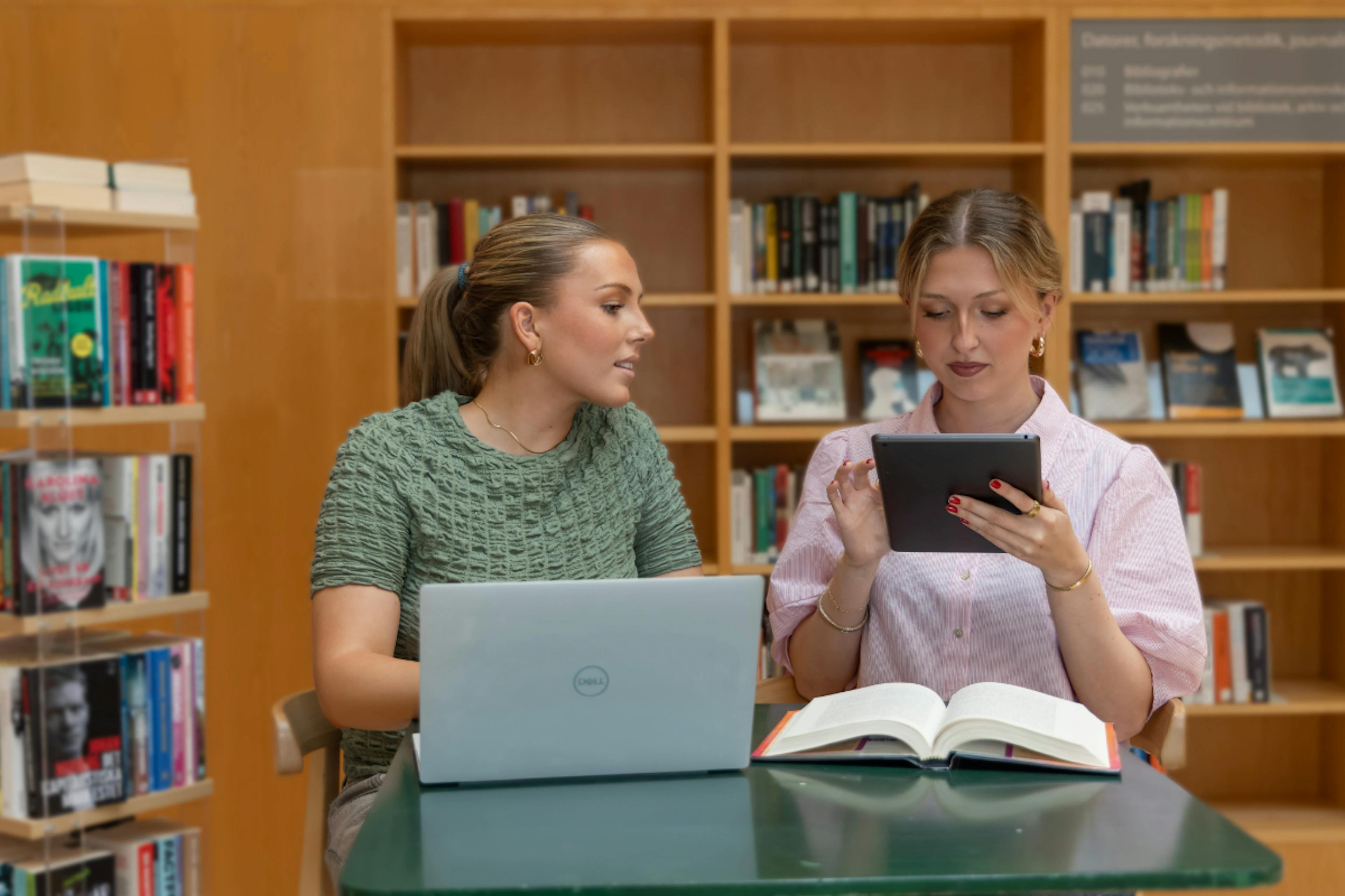 Två studenter i ett bibliotek som arbetar med en bärbar dator, surfplatta och en bok