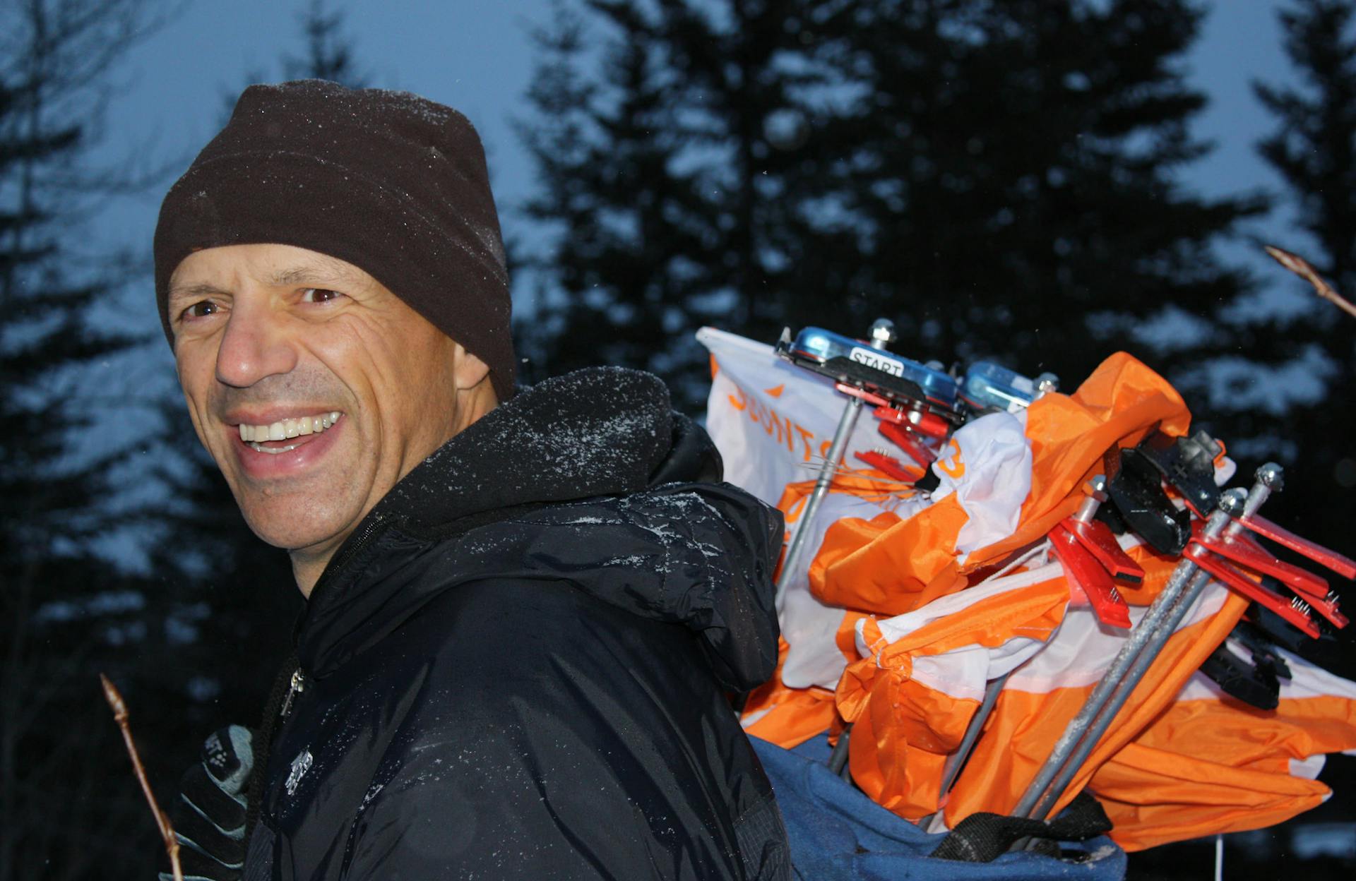 Adrian Zissos smiles at the camera while carrying a backpack full of orienteering controls. 