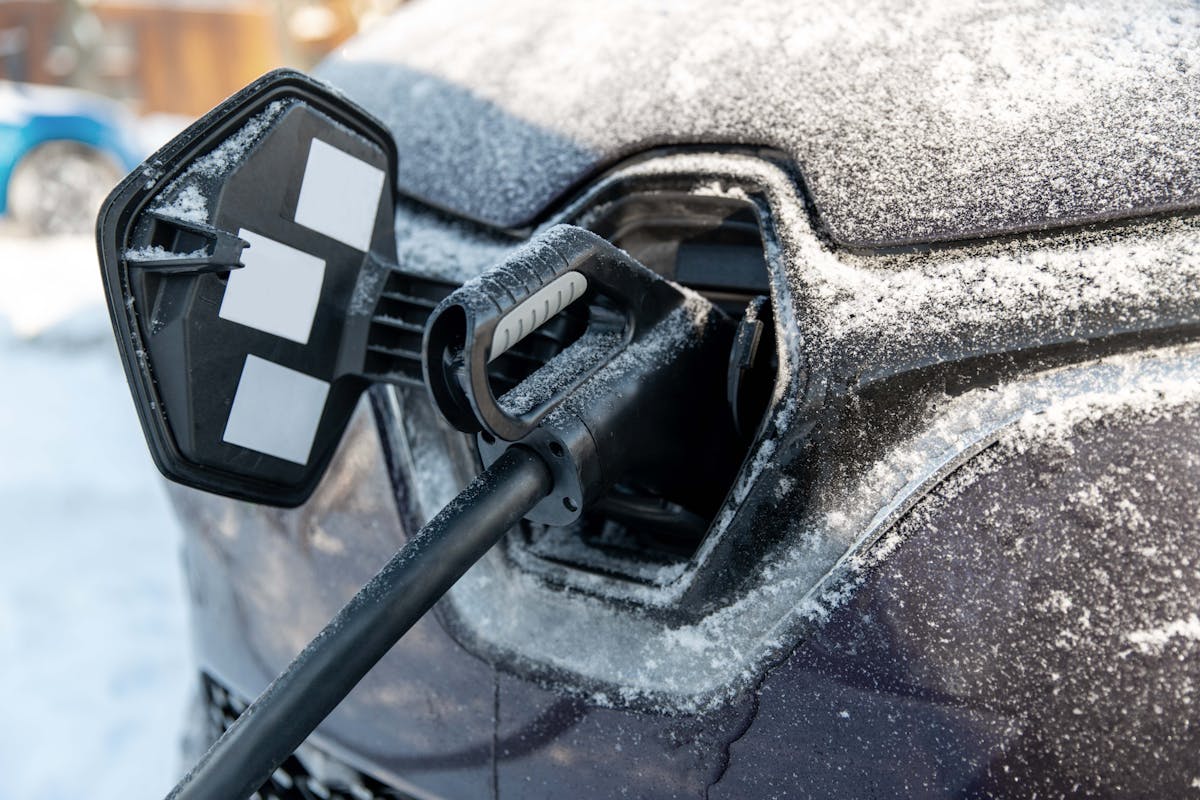 A black electric vehicle with a rapid charging cable plugged into its charge port. The car is covered in snow.