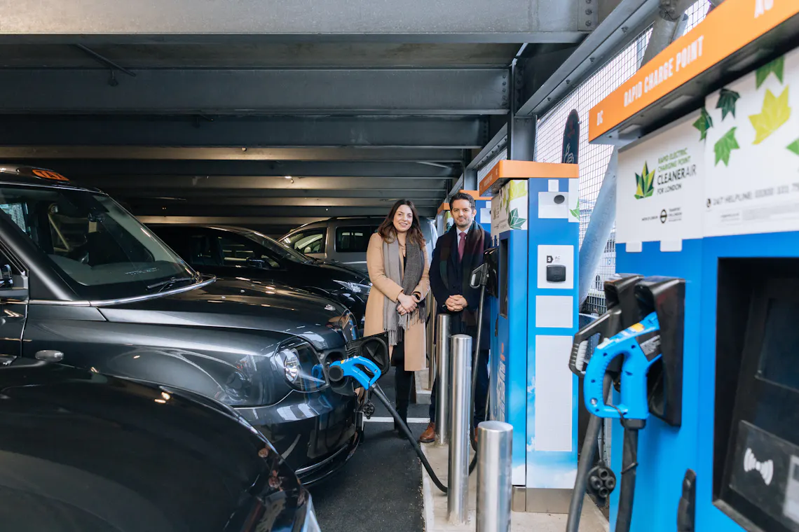 A man and woman stand beside an Osprey rapid charging hub while an electric taxi charges