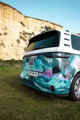 An electric VW ID Buzz van parked on the grass below a cliff.