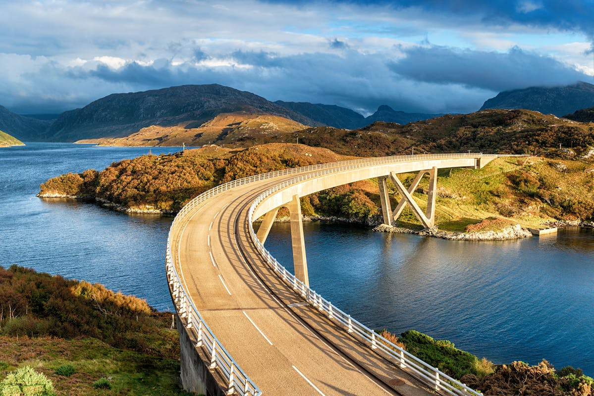 The curved Kylesku Bridge which crosses a lake in the Highlands of Scotland surrounded by hills covered in green and brown bush