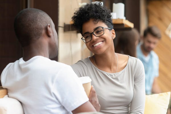 A young woman smiles at a young man sitting opposite her holding a cup of coffee.