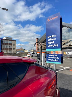 A red electric car parked in front of a sign for Electric Vehicle Rapid Charging in a retail park