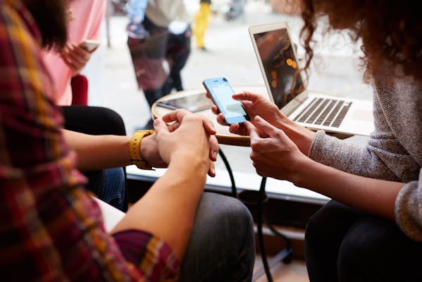 A young couple sitting at a table with a laptop on it, looking at the screen of a smartphone.