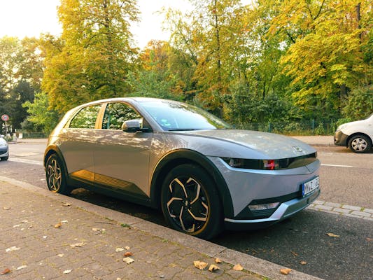 IONIQ 5 by Hyundai, a sleek electric vehicle, parked in the city centre with a striking grey metallic finish, surrounded by lush green trees.
