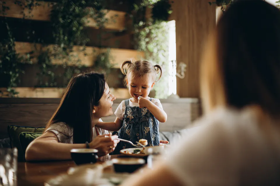 A toddler is smiling and standing at a table, next to her mother who smiles at her