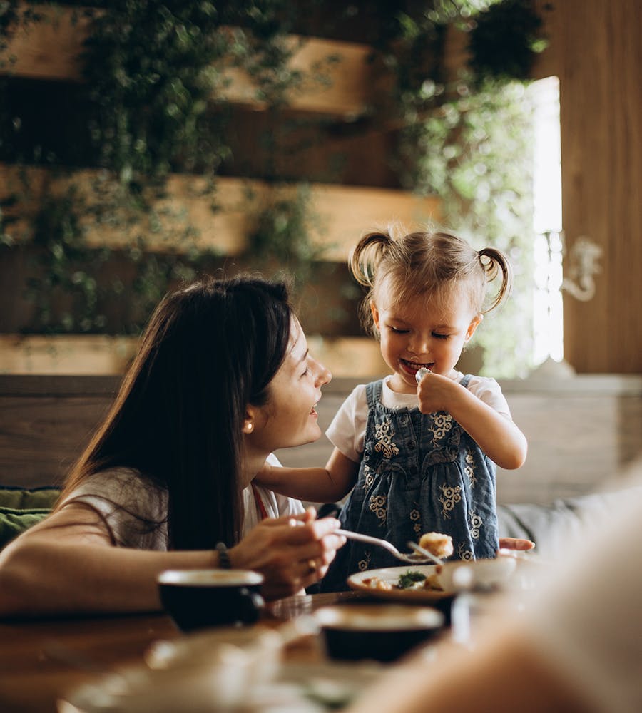 A toddler is smiling and standing at a table, next to her mother who smiles at her