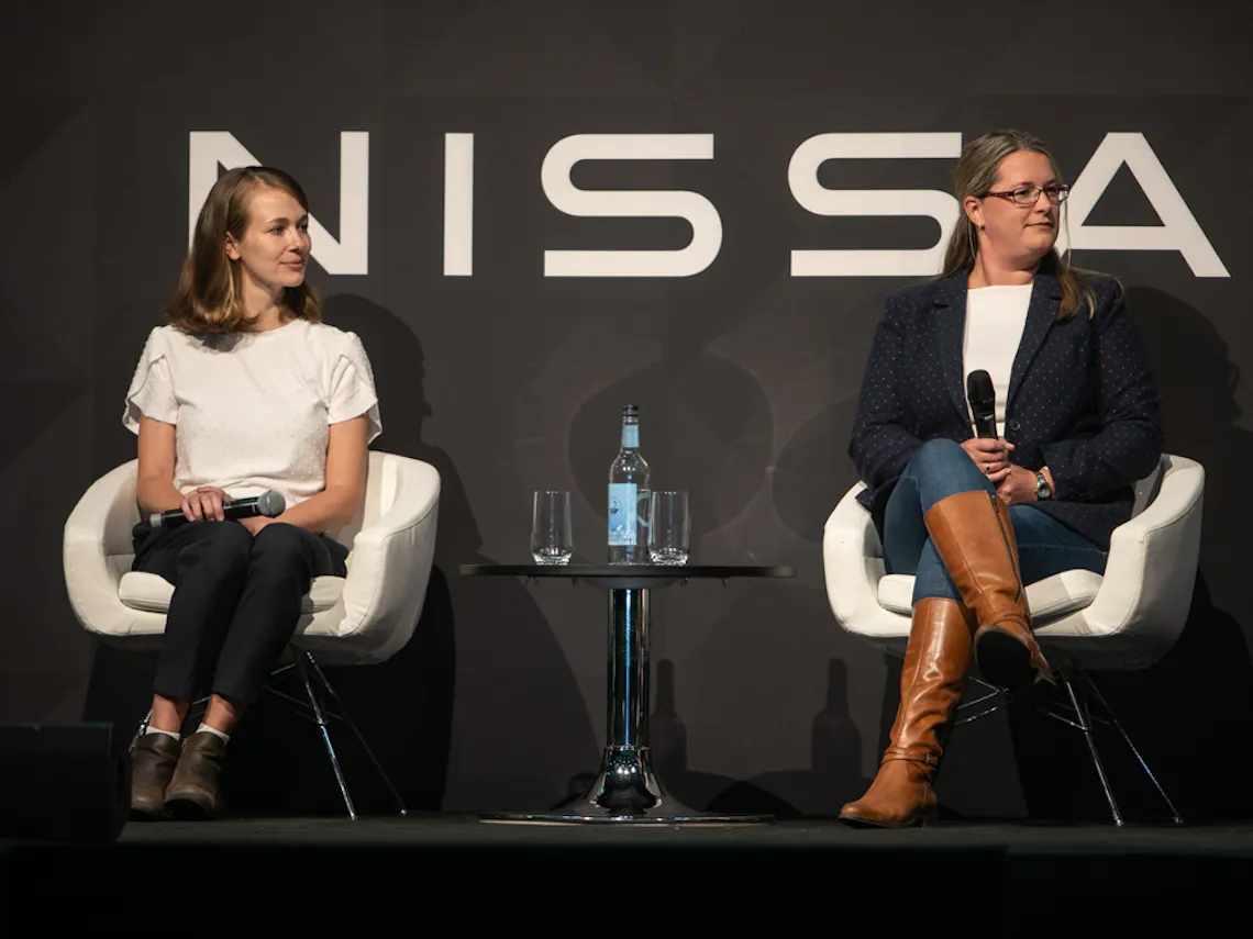 Two women sitting in white chairs on stage, holding microphones.