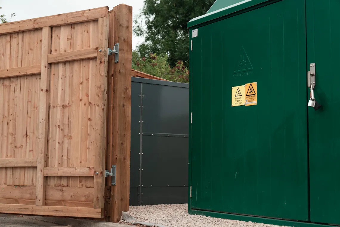 A large green substation behind a wooden fence