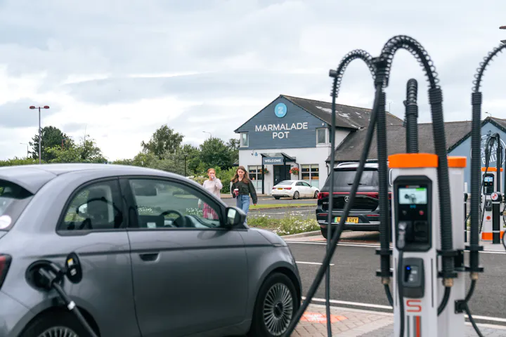 Two women with coffee walking towards an electric vehicle that is plugged into a high powered charger. 