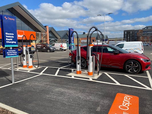 A red Kia EV6 charging at a four-charger rapid Osprey EV charging hub in a retail park with B&Q and M&S Food