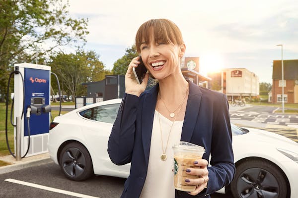 A woman talks on her smartphone while holding a Starbucks iced coffee, standing in front of a white electric car charging at an Osprey charging station.