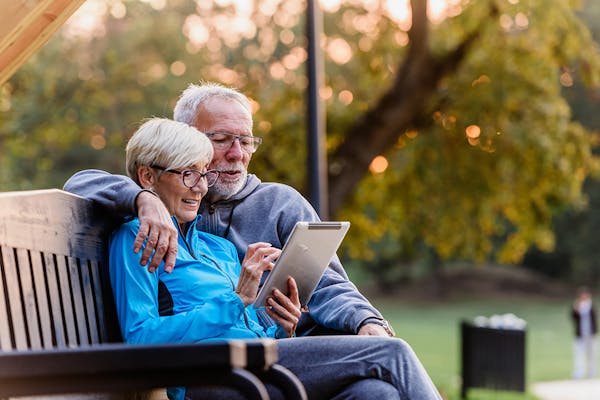 An older couple sit on a park bench together looking at a tablet