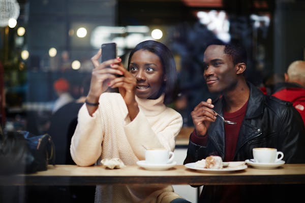 Two people sit drinking coffee and looking at a phone that one of them is holding up.