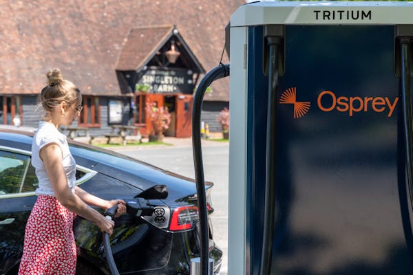 A woman in a red skirt and white blouse plugs her black Tesla into an Osprey EV charging station