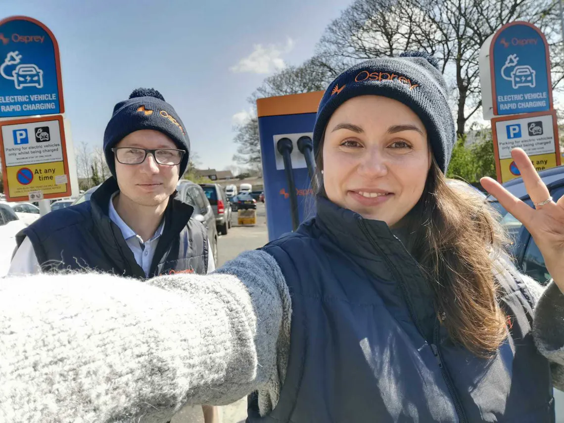 Two people wearing Osprey beanies pose in front of EV charge points