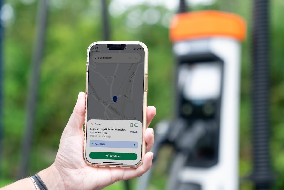 A hand holds a mobile phone with the Motability Go Charge app open in front of an Osprey rapid charging station. 