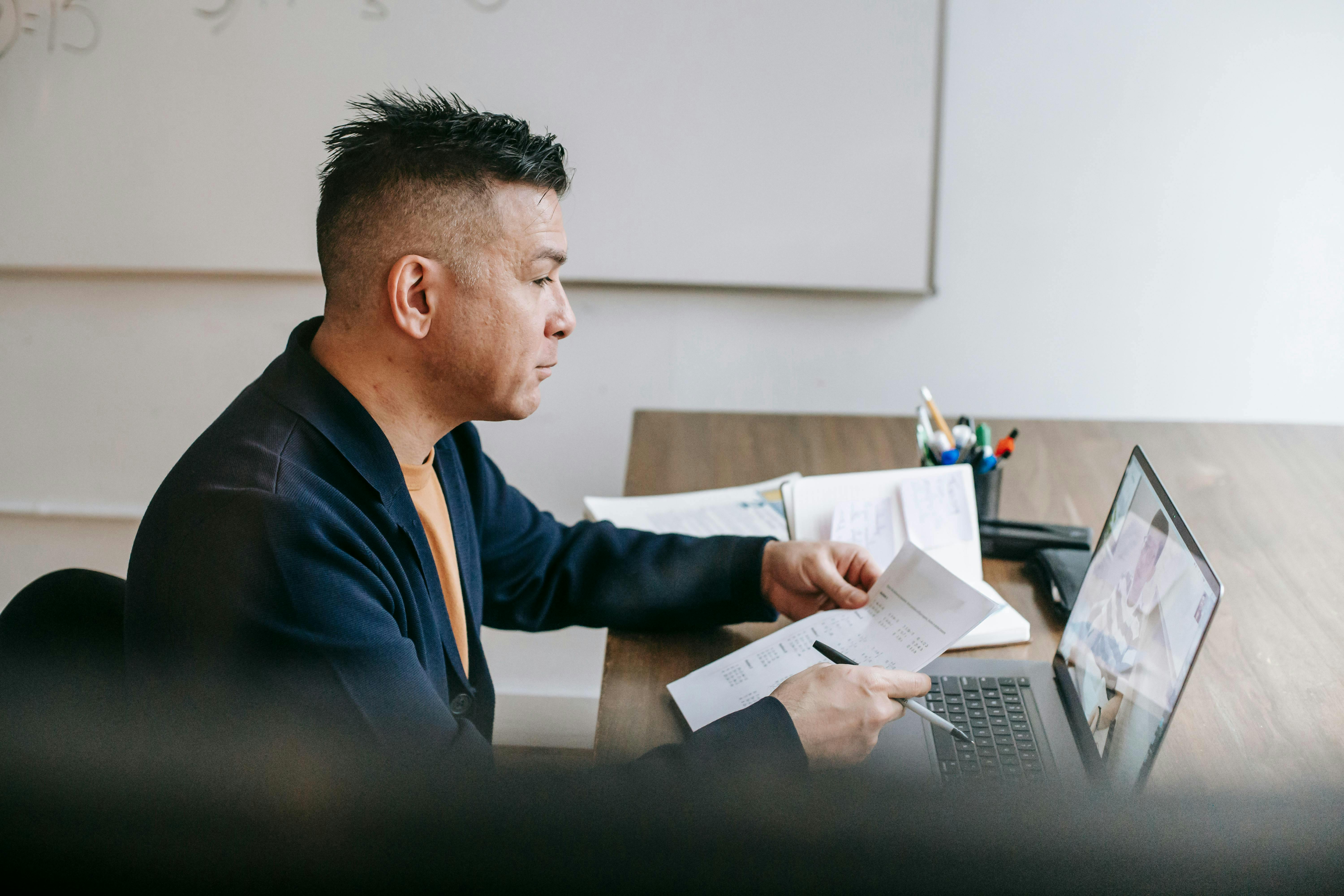 A man sits at a desk with a laptop, he is taking part in an online meeting. In his hand he holds a document and a pen.