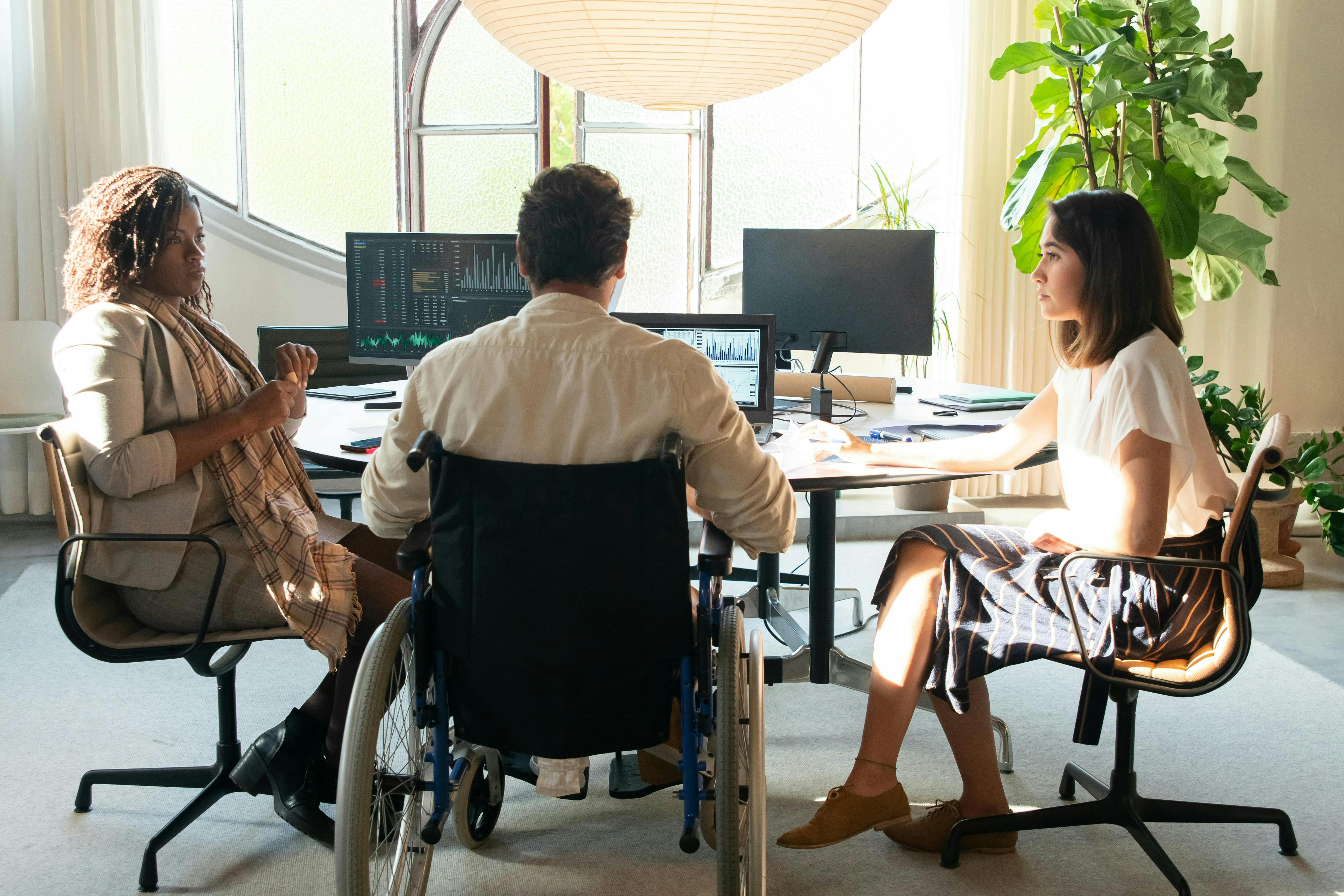 A group of three individuals of diverse identity, one in a wheelchair, sit working together at an office desk.