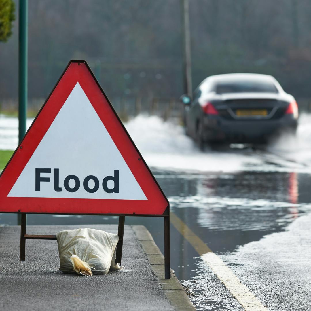 A triangular flood warning sign next to a puddle with a car driving through