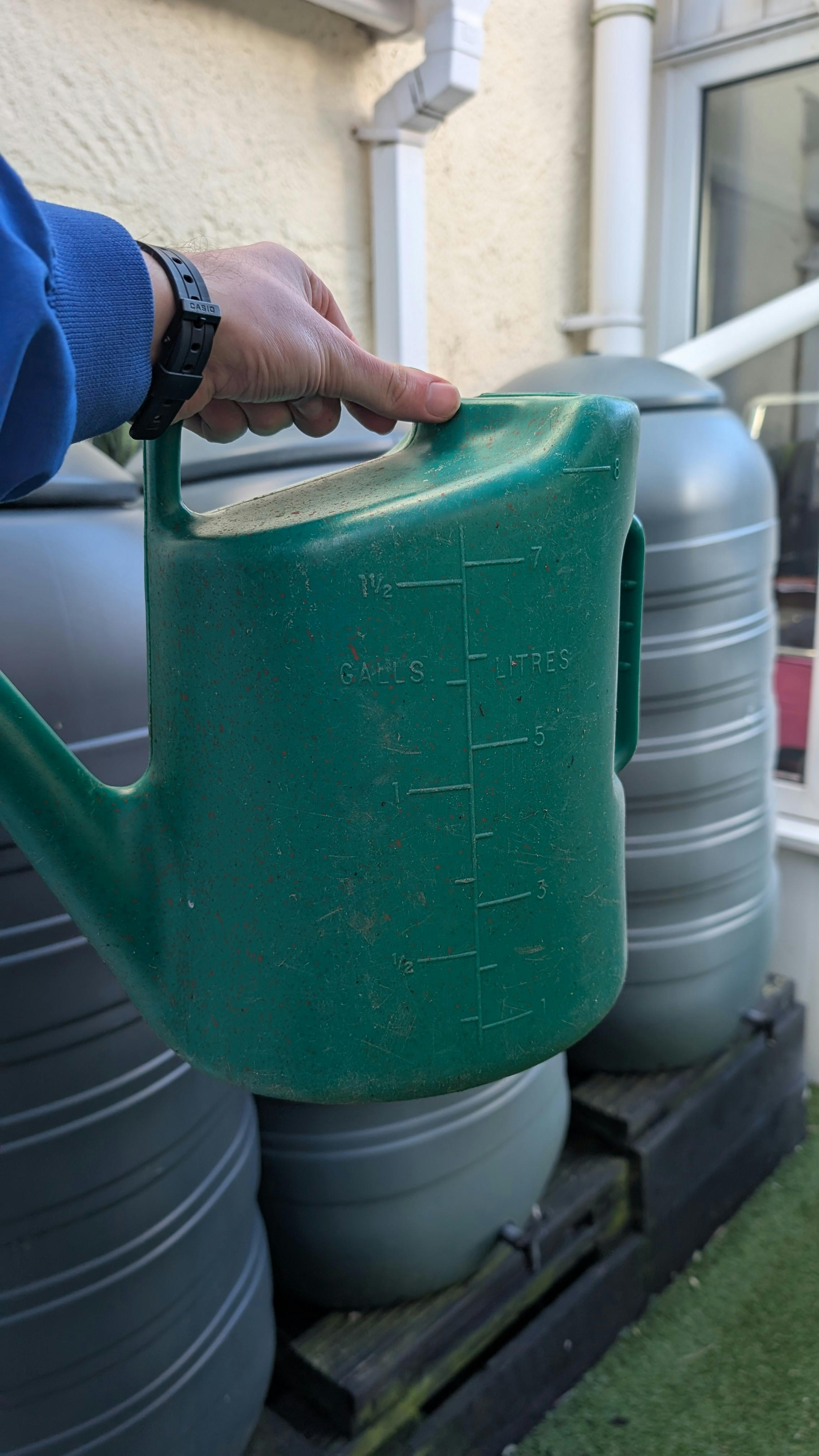 Green watering can being held up in front of three water butts.