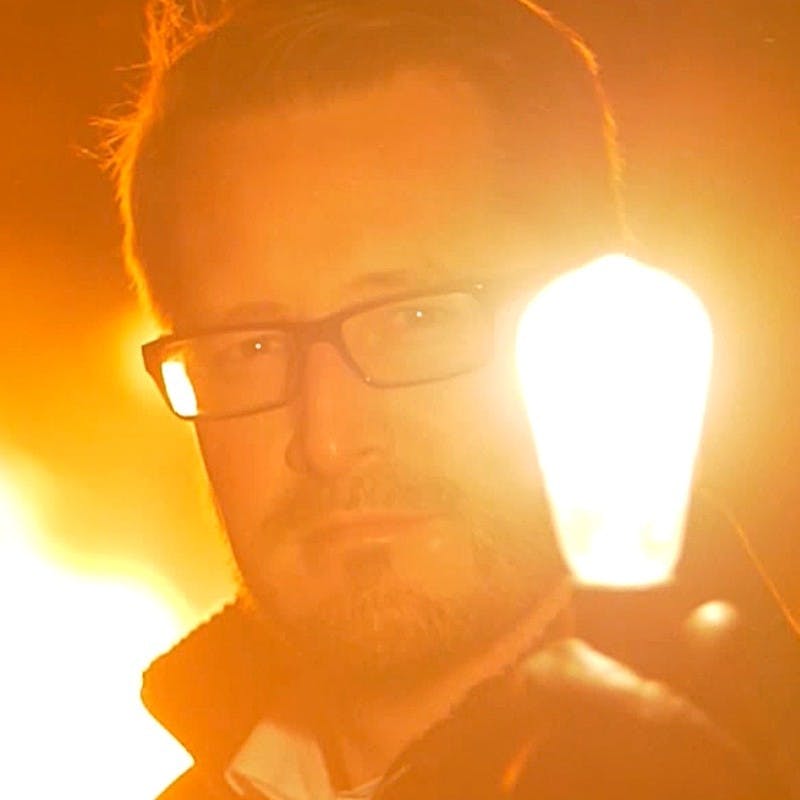A light-skinned man wearing glasses holding an illuminated lightbulb.