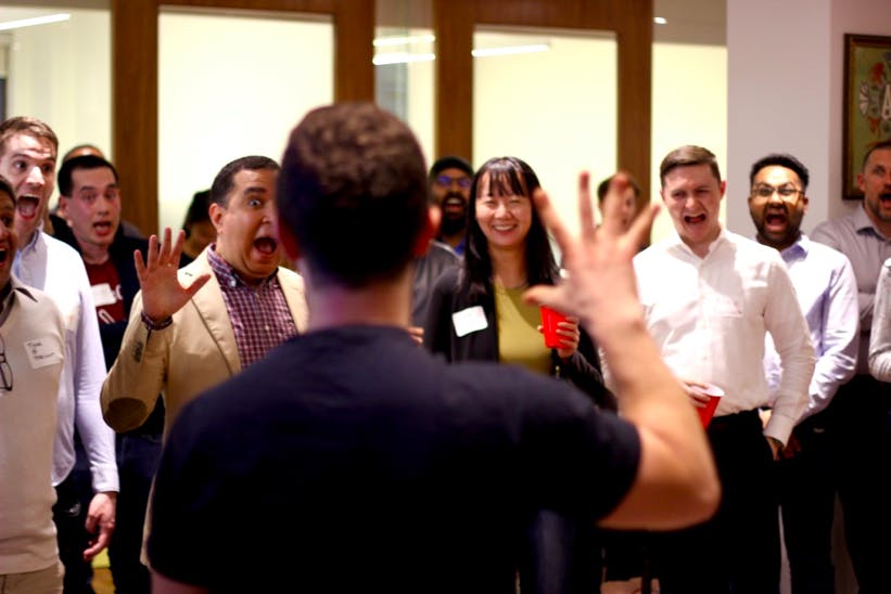 The back of a dark haired, light-skinned man speaking to a diverse group of people who watch attentively.