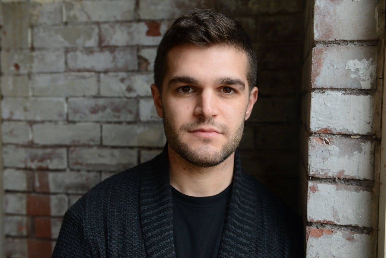 A light-skinned man with dark brown hair wearing a dark suit jacket and black t-shirt. He is standing in front of a painted brick wall.