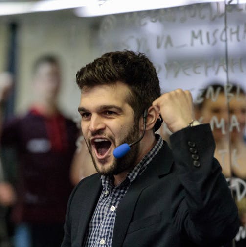 A dark haired, light-skinned man wearing a headset microphone pumps his fist in the hair while speaking to a crowd of people.