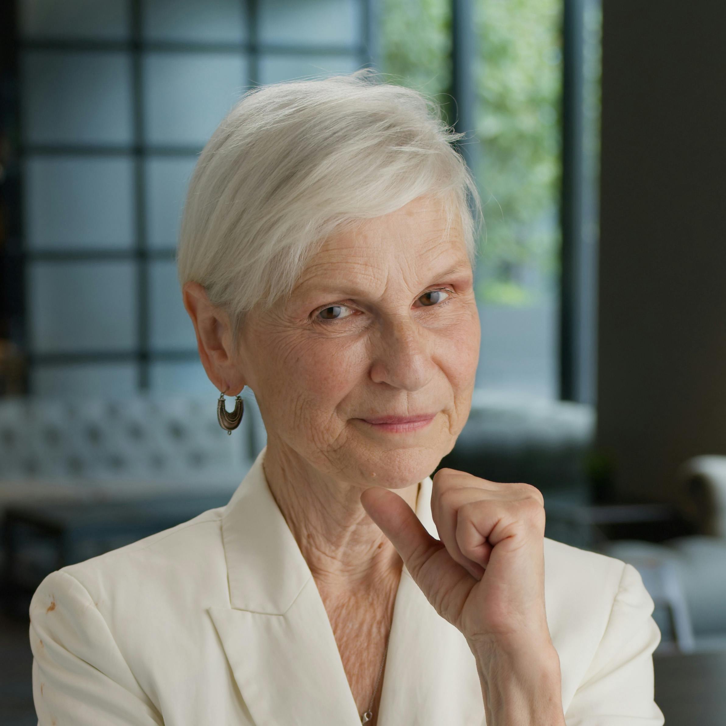 A light-skinned woman with short white hair wearing a white business suit jacket. She is looking into the camera with her head resting on her fist, with a window and sofa in the background.