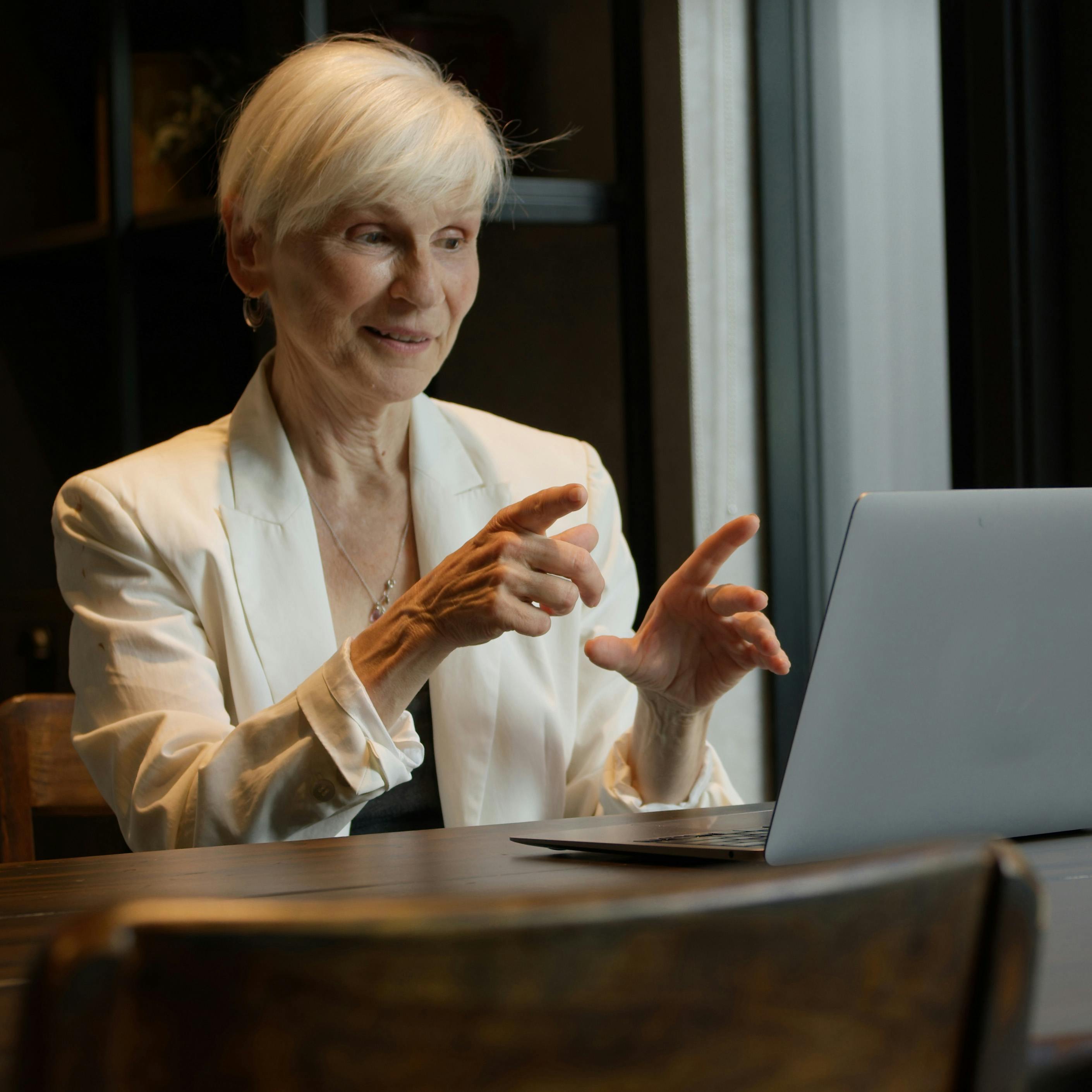A white haired, light-skinned woman in a white business jacket speaks into her laptop, gesturing with her hands.