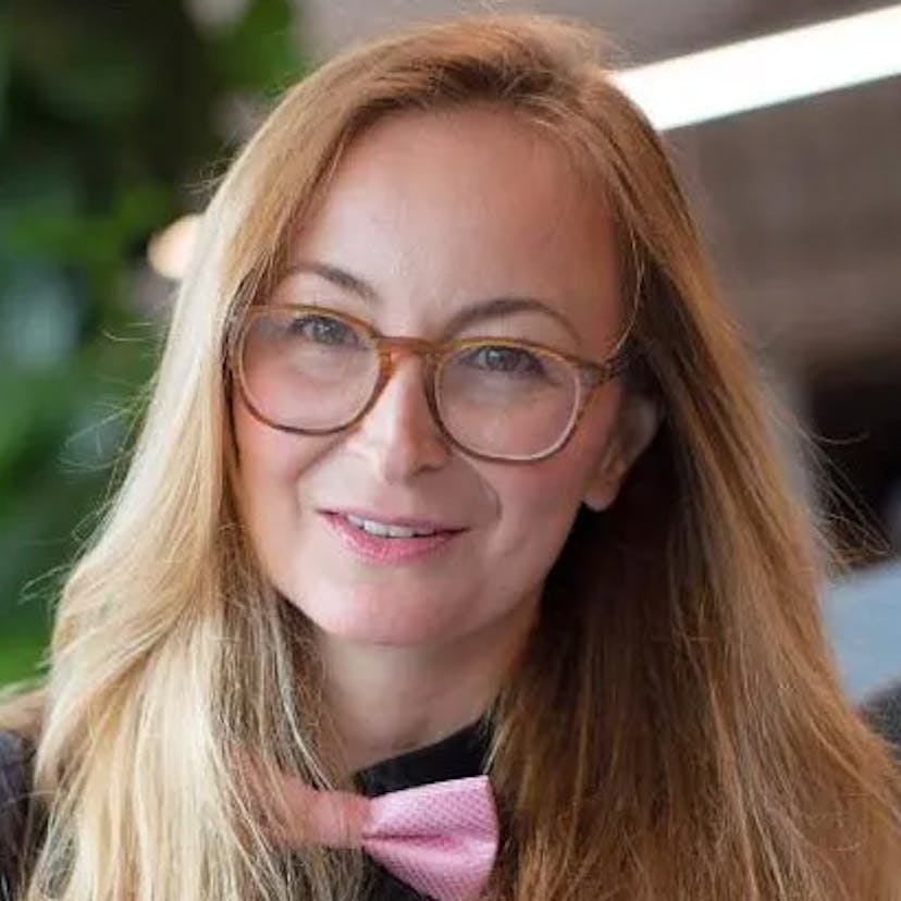A light-skinned woman with medium length blonde hair. She is wearing a pink bowtie and glasses and smiling slightly.
