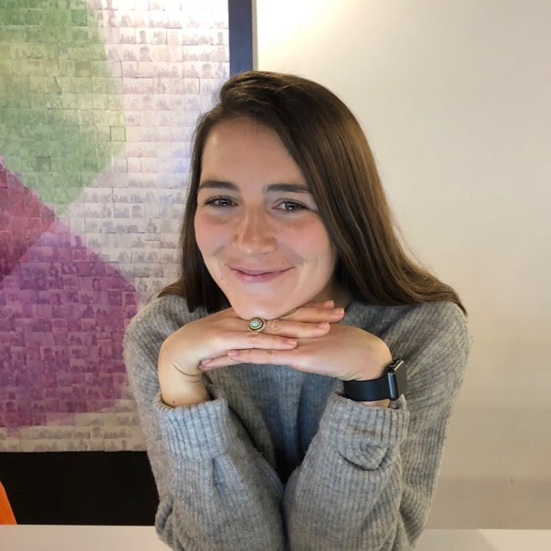 A light-skinned woman with long brown hair smiling while resting her head on her hands.