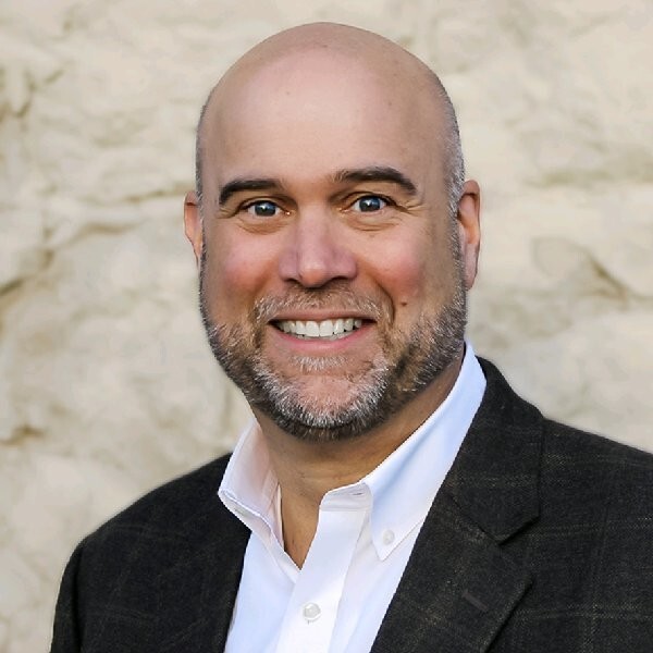 Steve Gielda wears a white collared suit and black blazer against a beige textured background.