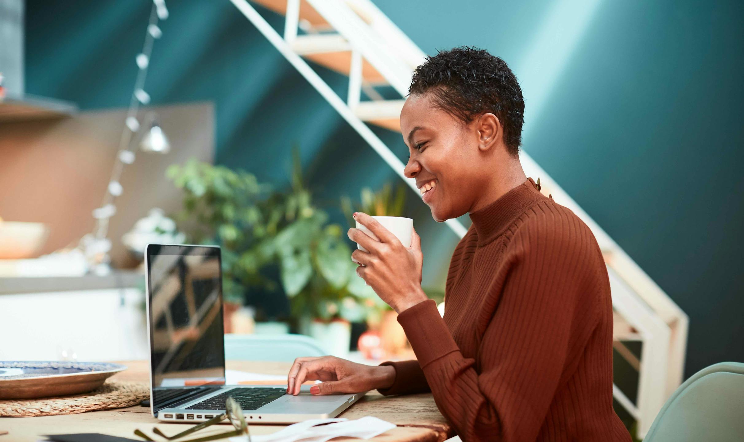 A Black woman holds a cup and smiles at her laptop as she sits in a sunlit office.