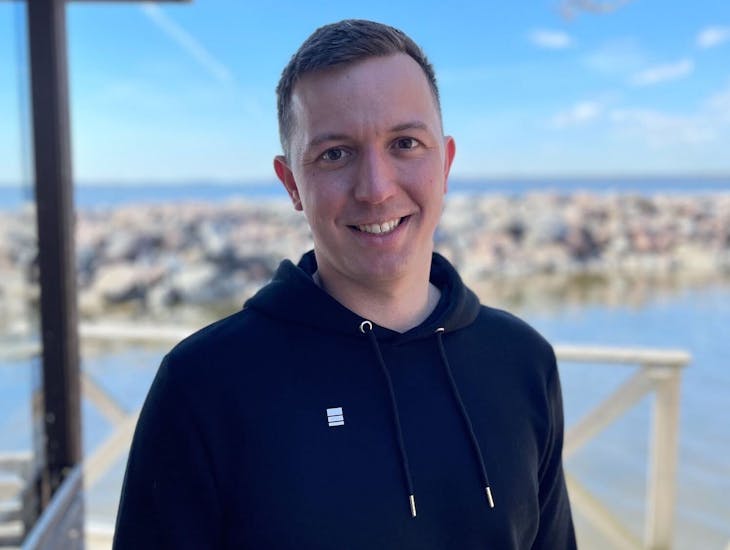 Epicbrief co-founder Anton Dobrzhanskiy smiles while wearing a black hoodie against a scenic coastline.