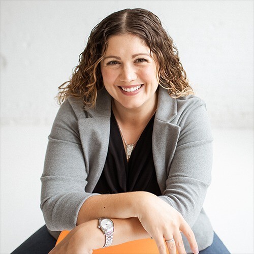 Kate DiLeo, founder of The Brand Trifecta, smiles at the camera with her wavy hair, her arms crossed, and a gray blazer on.
