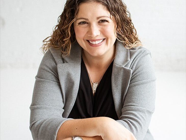 Kate DiLeo, founder of The Brand Trifecta, smiles at the camera with her wavy hair, her arms crossed, and a gray blazer on.