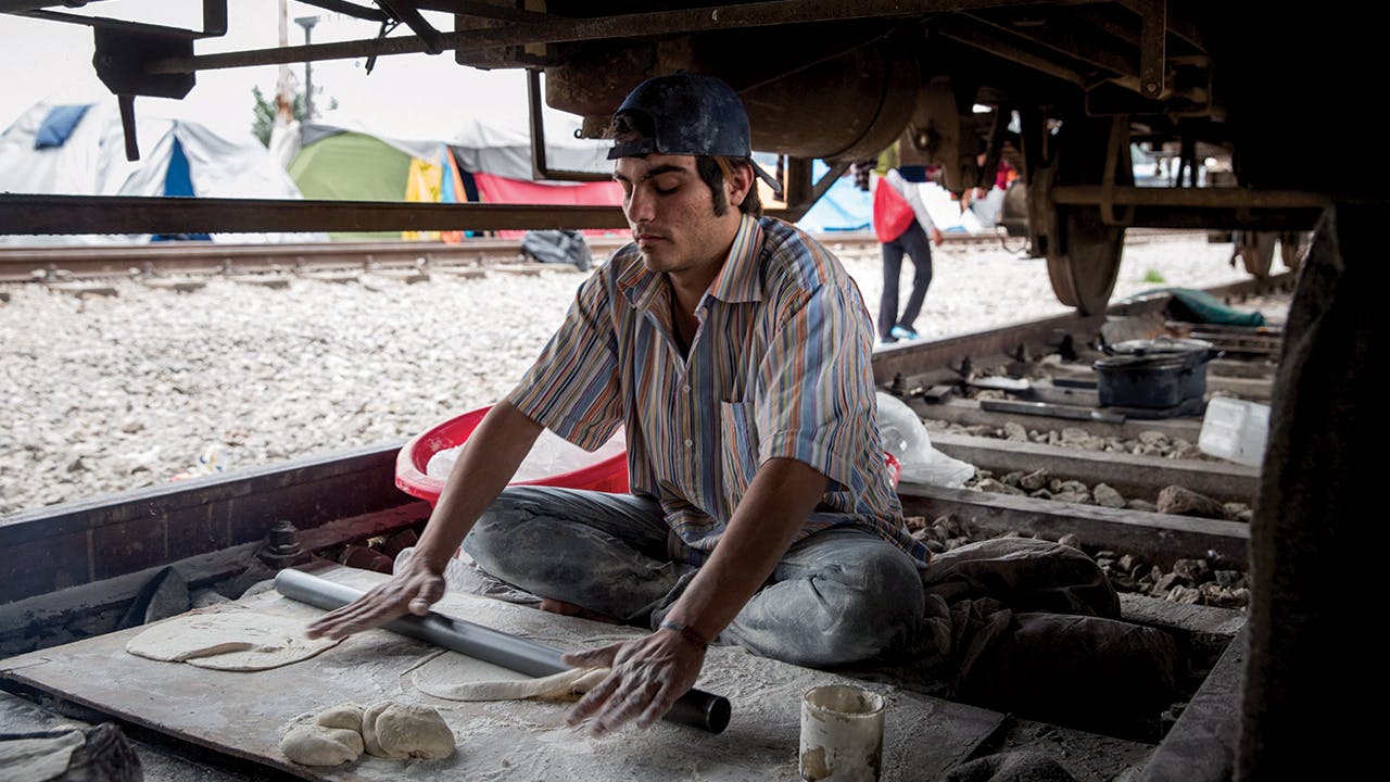 Makmoud Nakarch rolls out dough to make Syrian flatbread in his bakery under a cargo train parked at the Idomeni Camp in Greece. In 2016, after European borders were closed, more than 14,000 asylum-seekers were stranded, living in the area for months.