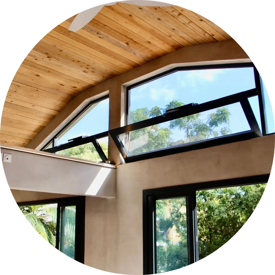 View from inside the house, with hinged windows on the upper part and a section of sliding windows below, overlooking the outside where dense vegetation and the blue sky can be seen.