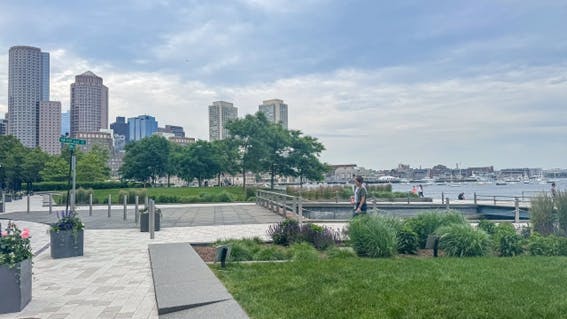 Fotografia de uma paisagem urbana. Ao fundo, há prédios. São observadas também árvores e espaços com gramado. Algumas pessoas caminham. No canto direito, aparece um lago.