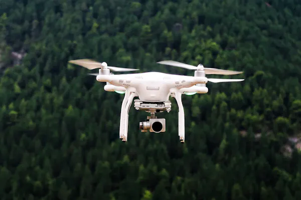 Posicionado no centro da fotografia, um drone com uma câmera instalada sobrevoa uma floresta.