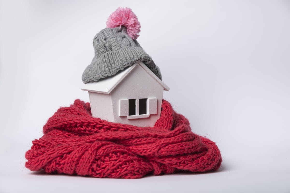 Model of a home, wrapped in a red scarf and wearing a gray hat with a pink puff ball, representing a home that's using winter energy saving tips to be more energy efficient.