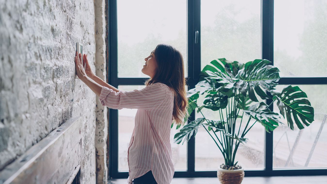 A woman adjusts a thermostat in a brightly lit room.